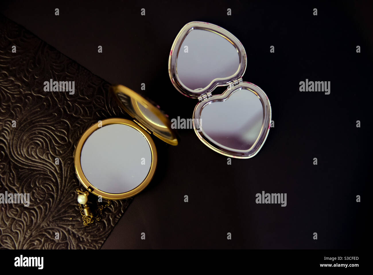 a contrast between a gold round pocket mirror and a silver heart-shaped pocket mirror on a half-textured black background - Smartphone Captured Stock Image