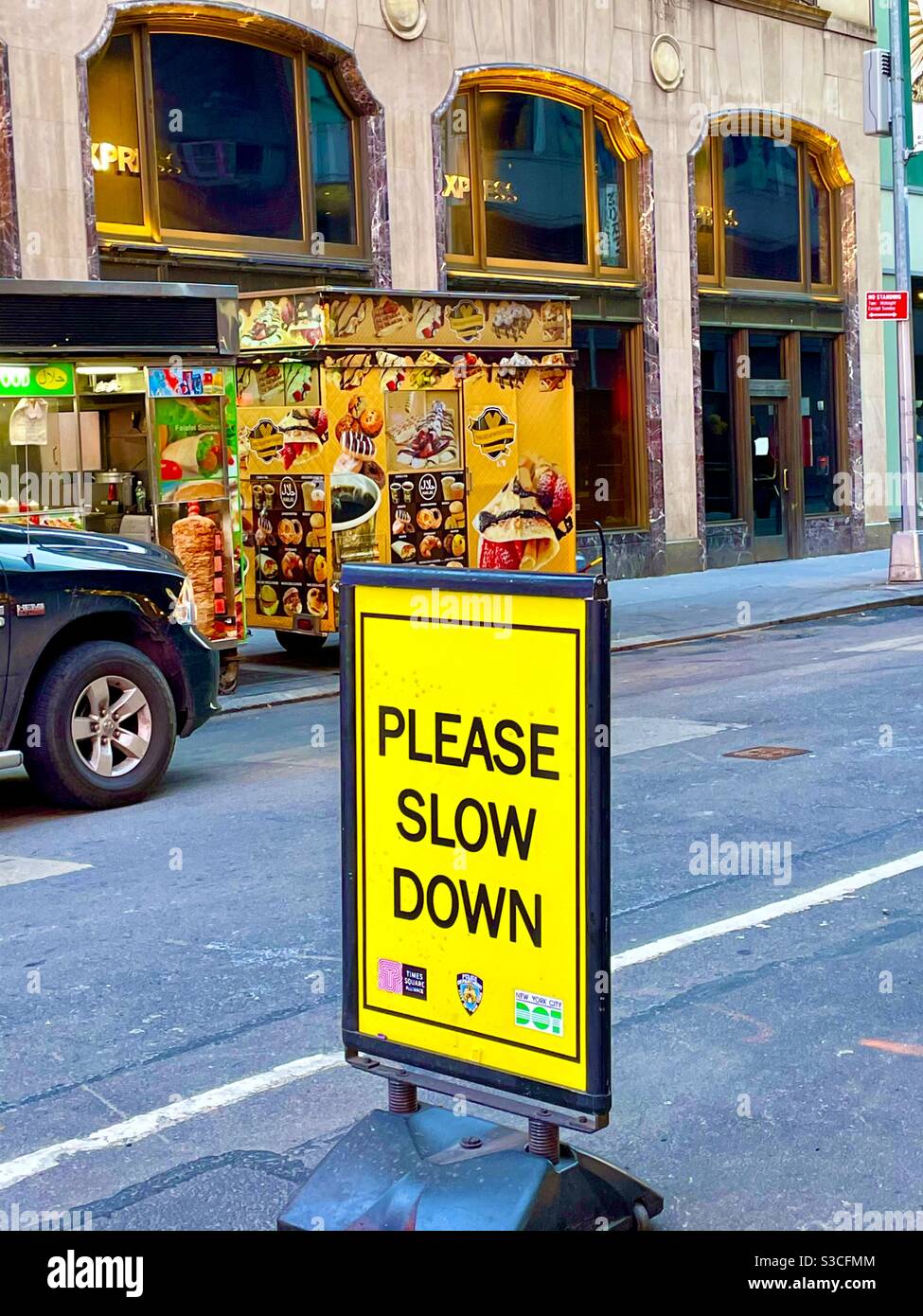 A simple bright yellow street sign contrasts with the busy multicolored signage of a street vendor in front of A beautiful architecturally embellished spaceBuilding in Times Square, Manhattan, USA. - Smartphone Captured Stock Image