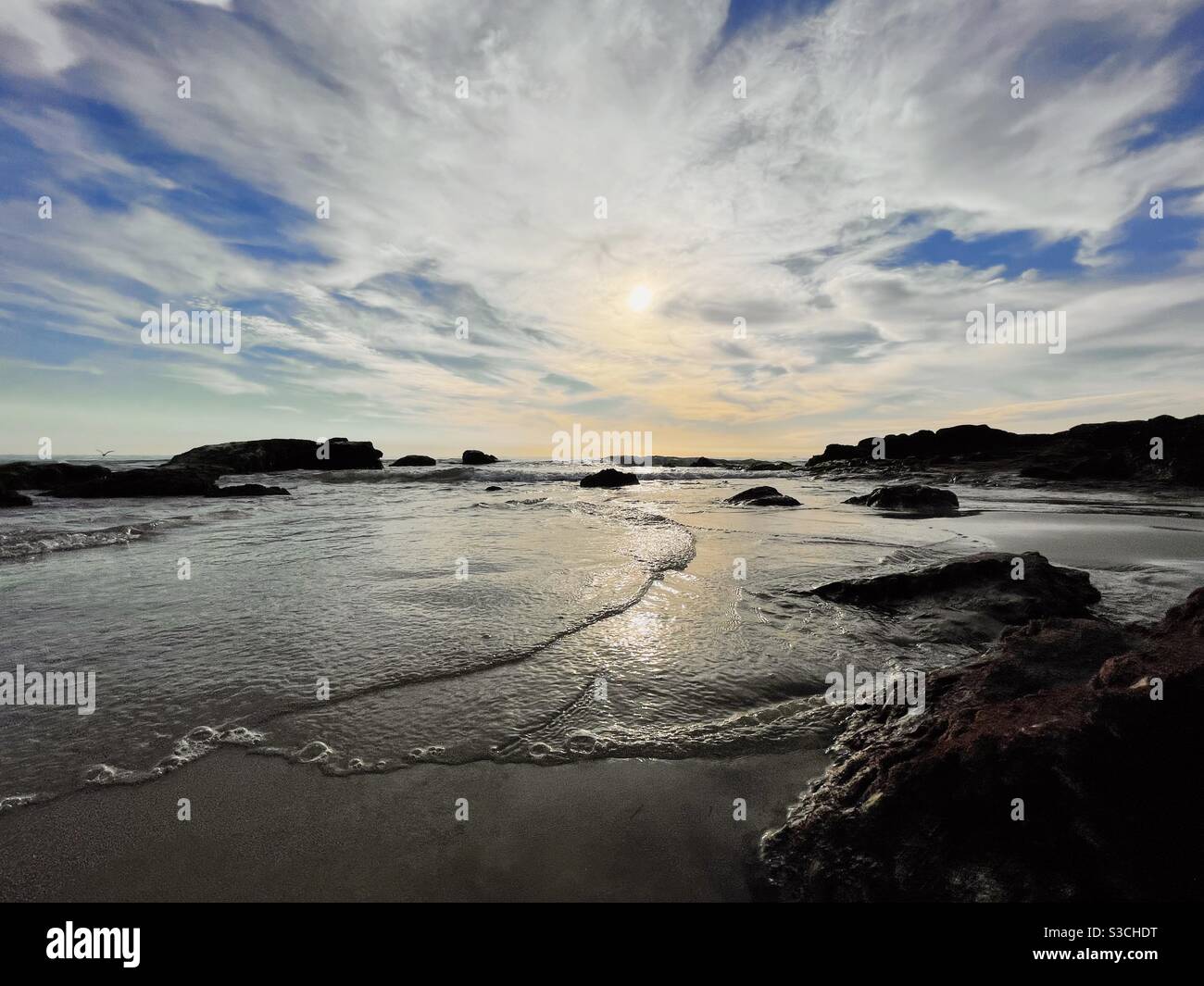 Late afternoon seascape with gentle waves washing up onto sandy beach amongst rocks with dramatic sky above. - Smartphone Captured Stock Image