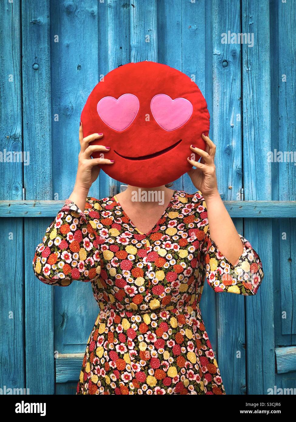 Woman in colorful dress standing against a blue wall and holding pillow with hearts in front of her face - social media concept for love - Smartphone Captured Stock Image