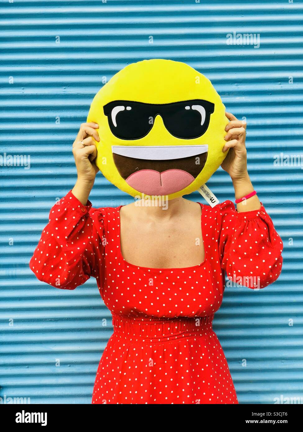 Woman standing against blue wall and holding a smiley face pillow over her face - social media concept for happiness - Smartphone Captured Stock Image