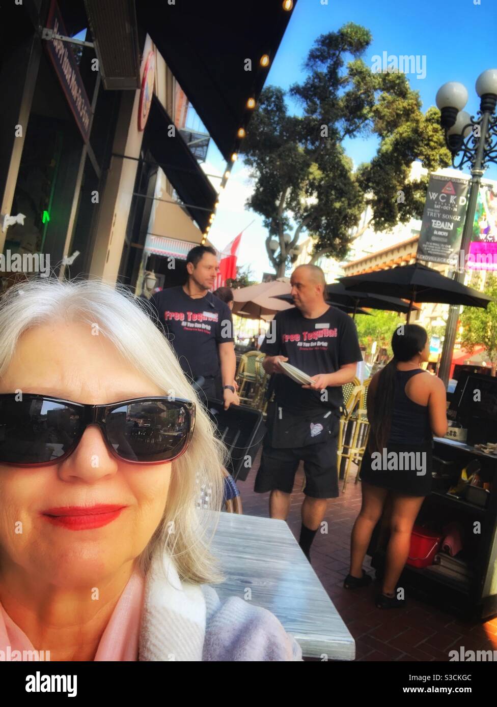 An American street scene in the Gaslamp district of downtown San Diego, California, USA where a woman in sunglasses sits at an outdoor café along a busy sidewalk - Smartphone Captured Stock Image