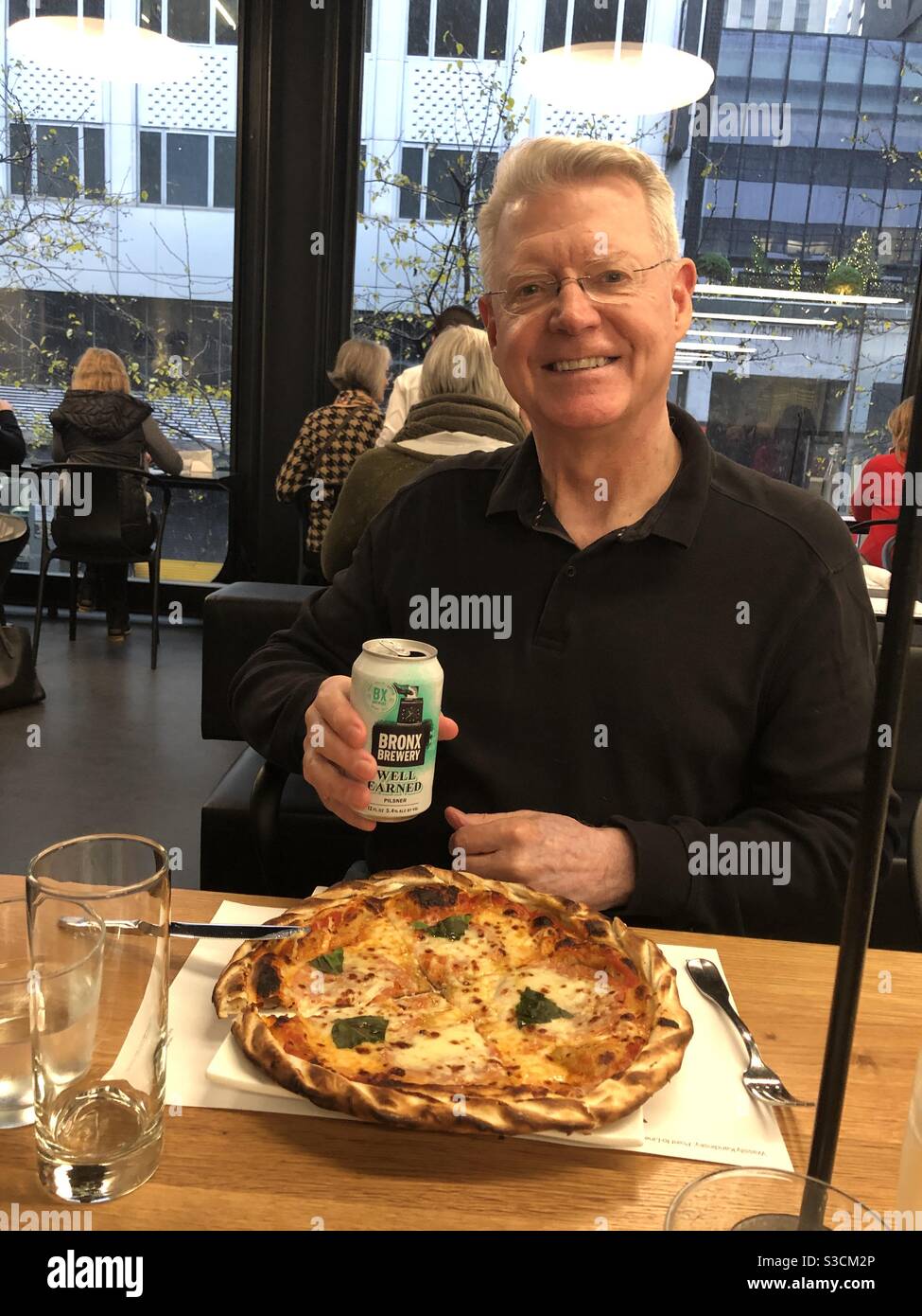 A serendipitous light reflection creates a halo over a smiling man eating pizza and beer in the cafe at the Museum of modern Art in New York City, USA; Does he deserve this Halo? - Smartphone Captured Stock Image