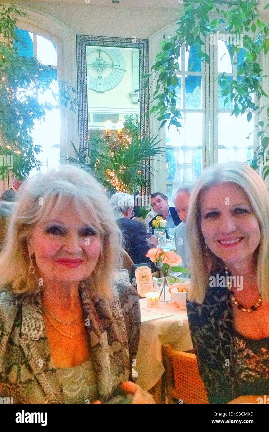 Two blonde woman, mother and daughter, share big smiles in a beautiful restaurant in studio city, Los Angeles, California with large gazebo windows dripping with plants - Smartphone Captured Stock Image