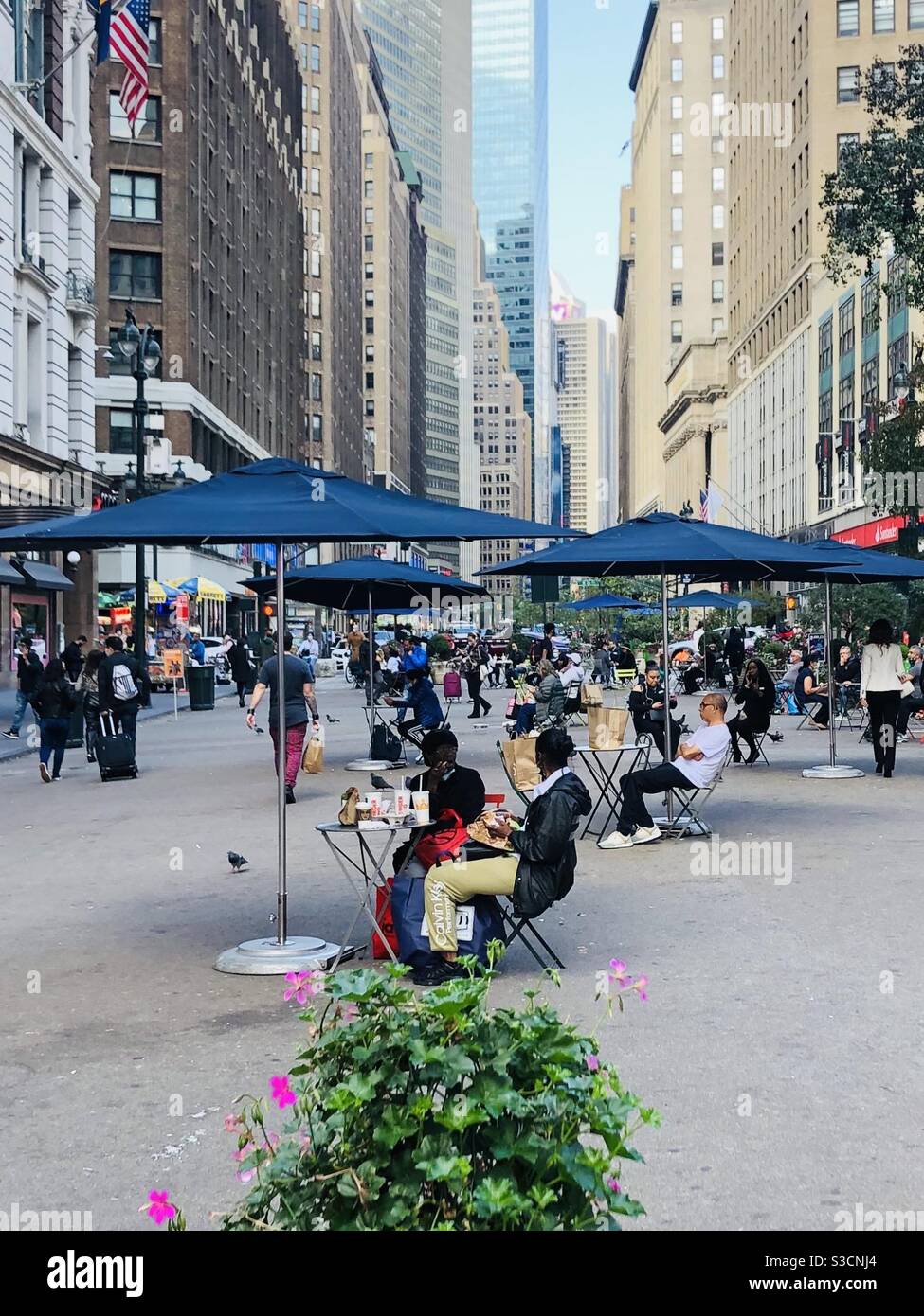 A painter leaving yet of New York City life on the sidewalks of Herald Square in Midtown Manhattan as people stroll or sit under blue umbrellas surrounded by skyscrapers on a quiet afternoon - Smartphone Captured Stock Image