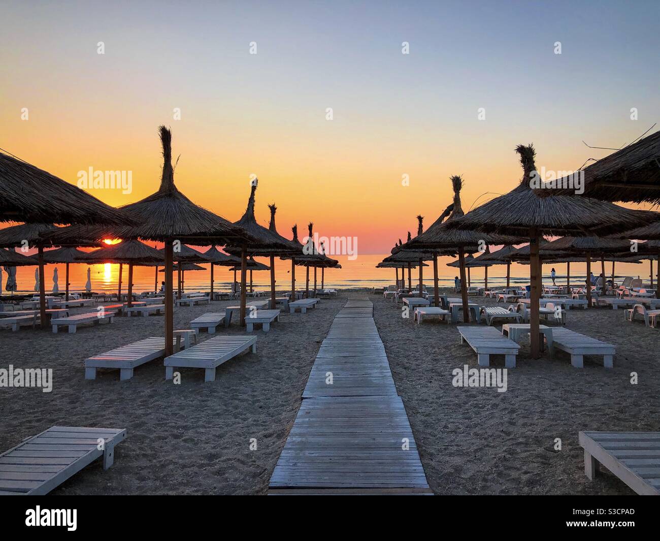 Thatched roofs umbrellas and sunbeds on an empty beach at sunrise - Smartphone Captured Stock Image