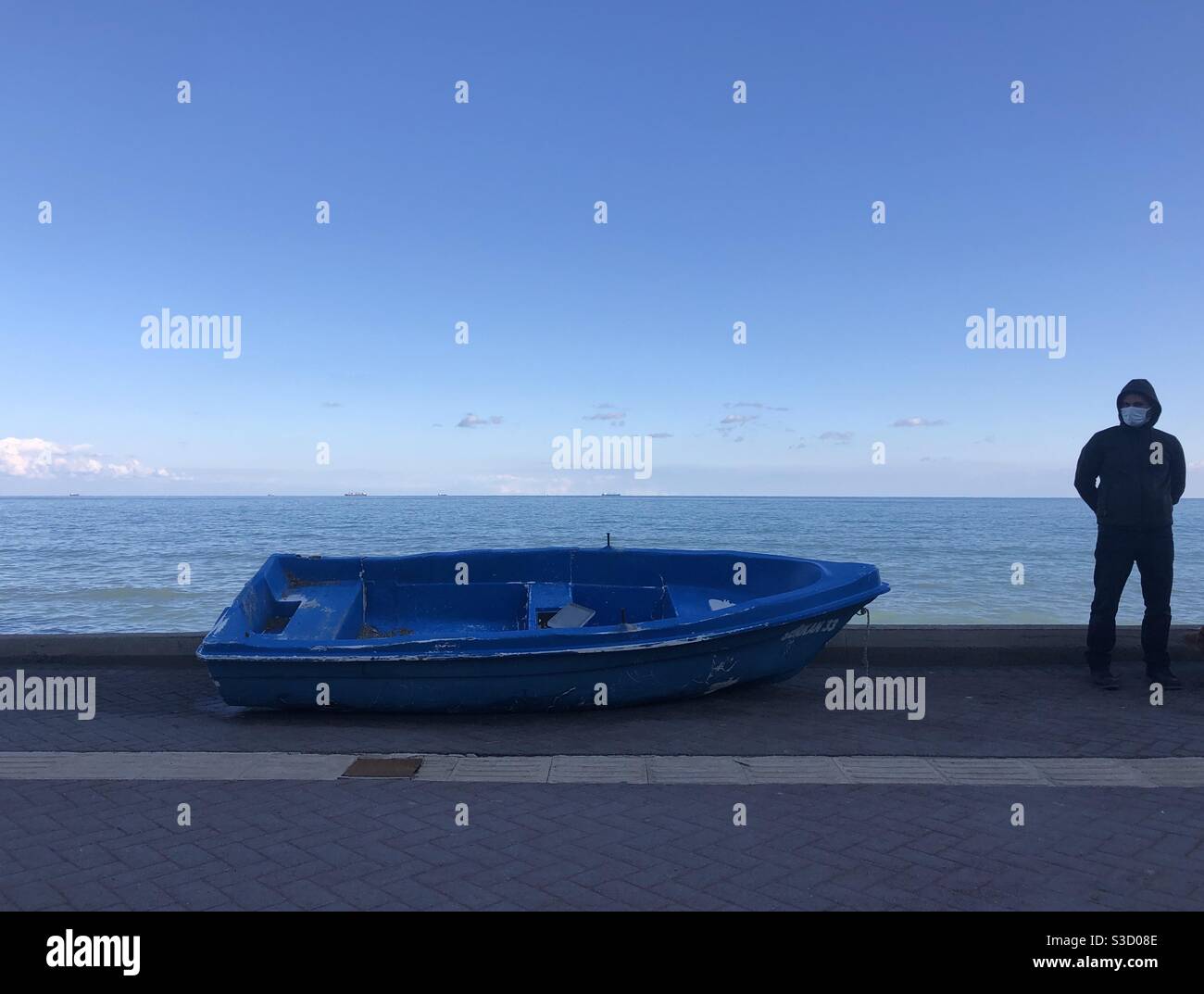 Man wearing a face mask standing next to a blue fishing boat by the sea - Smartphone Captured Stock Image