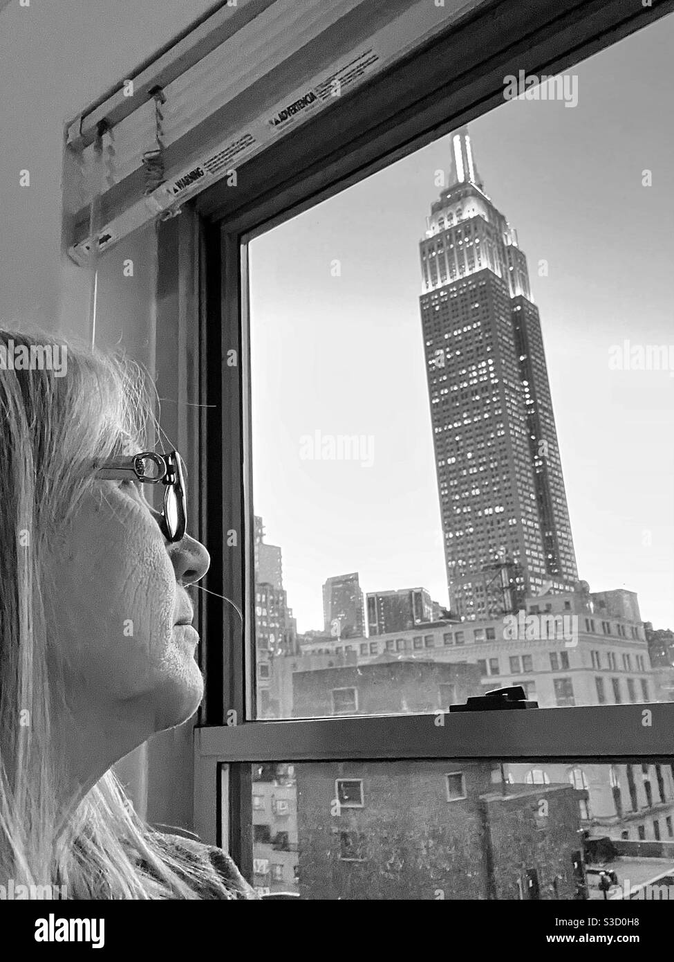 What is this woman thinking as she gazes out a New York City apartment window at the Empire State Building standing out like a sentinel among skyscrapers?? - Smartphone Captured Stock Image