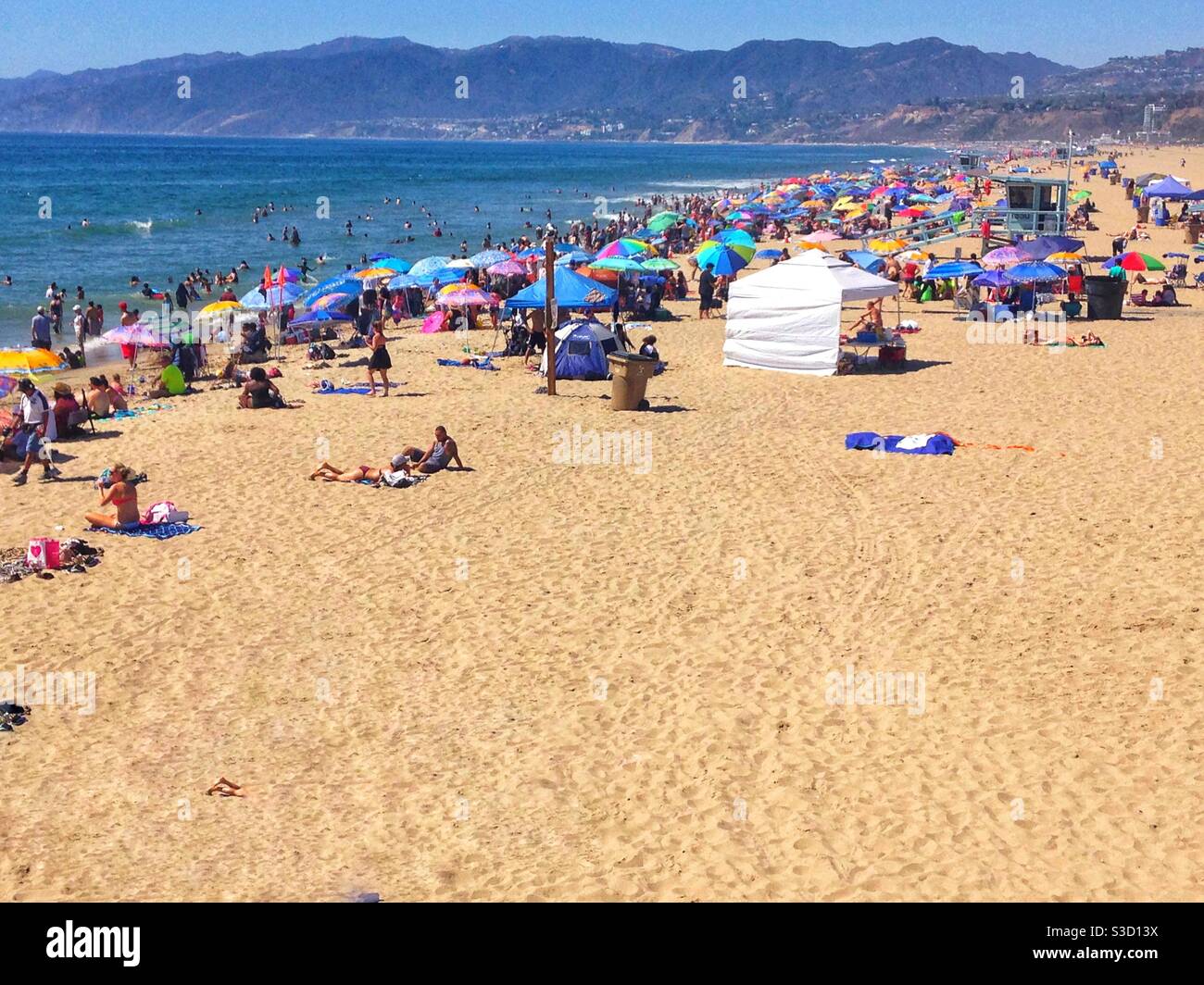 Crowds of people, some under colorful umbrellas, enjoy a sunny afternoon at the beach in Santa Monica, Los Angeles, California, USA with a view of Malibu in the background - Smartphone Captured Stock Image