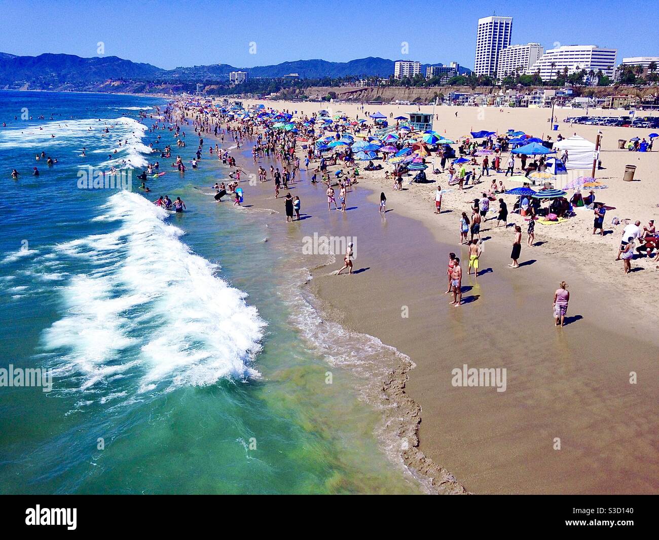 A glorious day to be at the beach! People, pets, surfers, colorful umbrellas and a view of buildings in Santa Monica as well as the hillsides of Malibu make this scene idyllic! Stock Photo