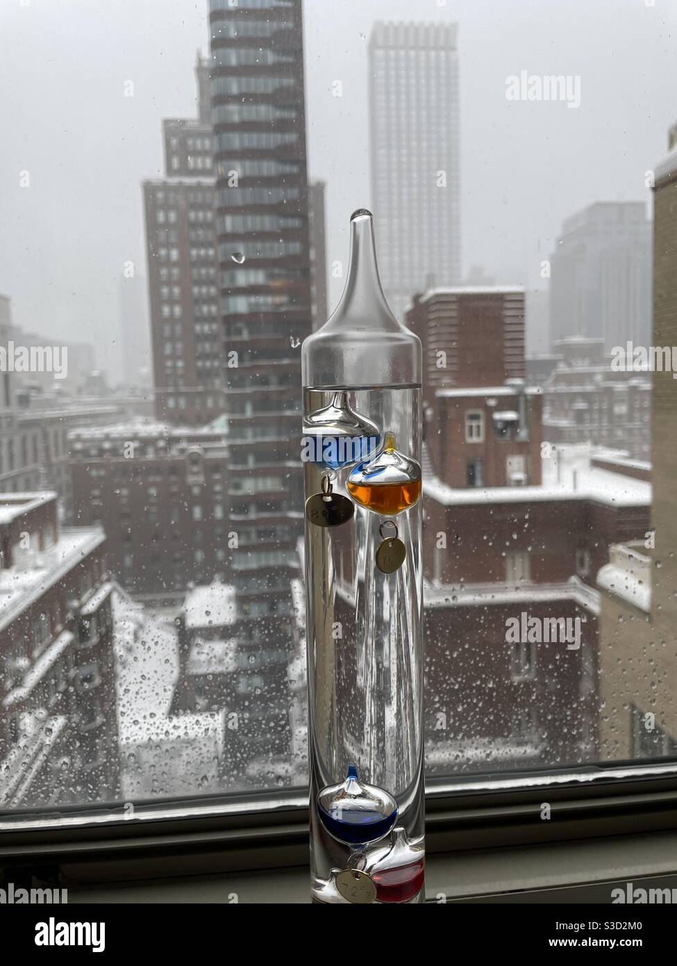 February 1, 2021 blizzard, a Galileo thermometer in a window viewing snow covered terraces and skyscrapers obscured by snowy skies in Manhattan, New York, USA - Smartphone Captured Stock Image
