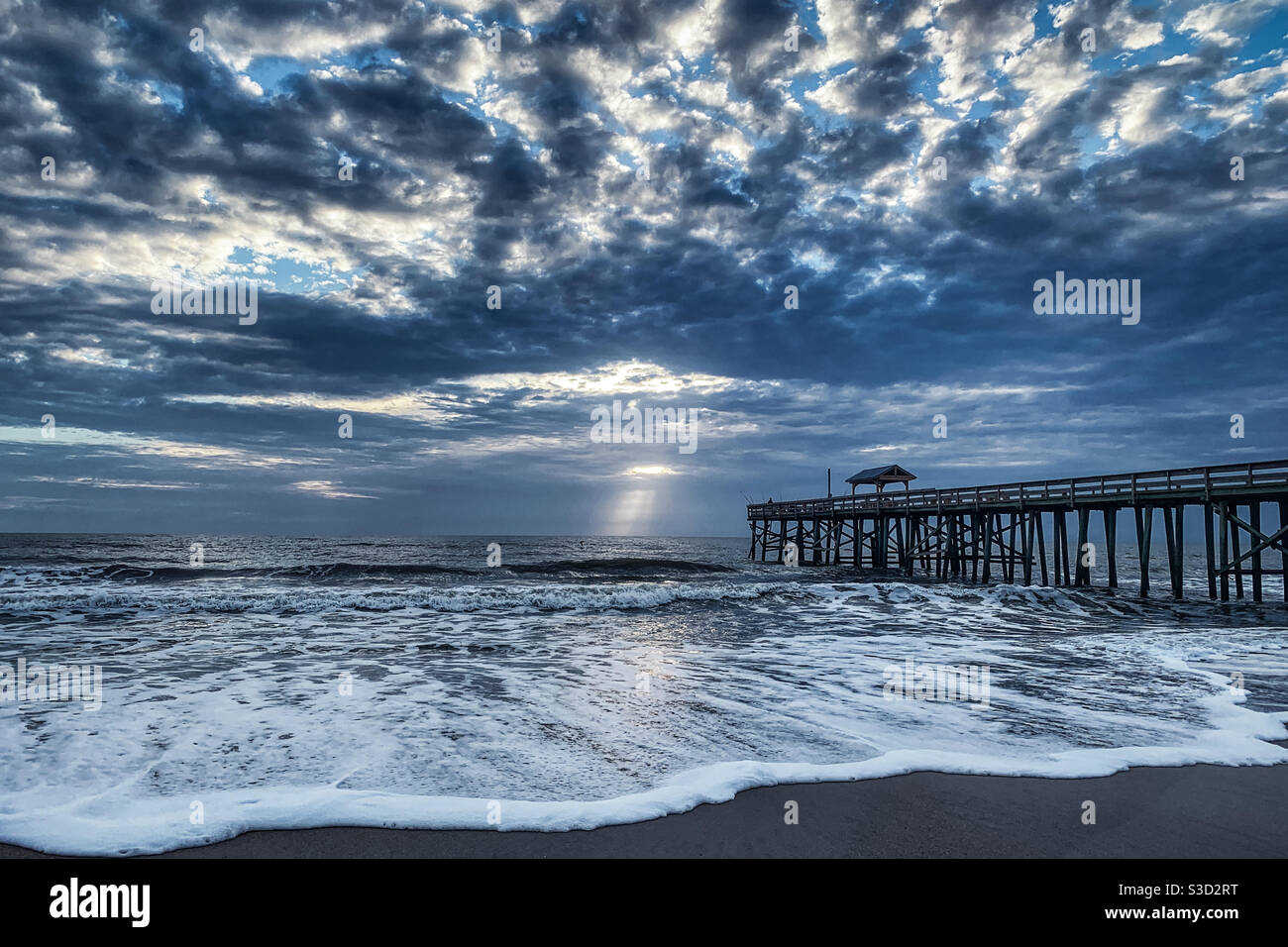 Crepuscular rays shining through the clouds over the Amelia Island beach pier with ocean waves rolling in. Stock Photo