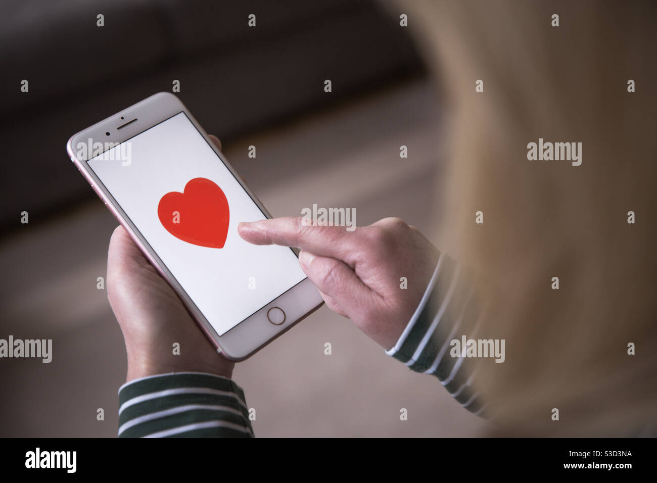 Hey girl holding a mobile phone and tapping on a love heart in a romantic message or dating app - Smartphone Captured Stock Image