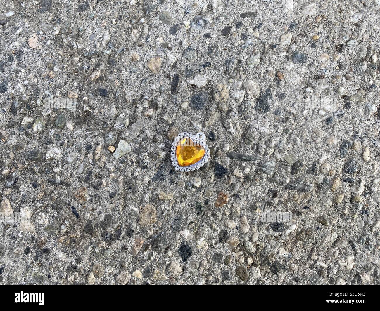 Plastic amber, heart-shaped costume jewelry trinket that's been dropped on a stone sidewalk - Smartphone Captured Stock Image