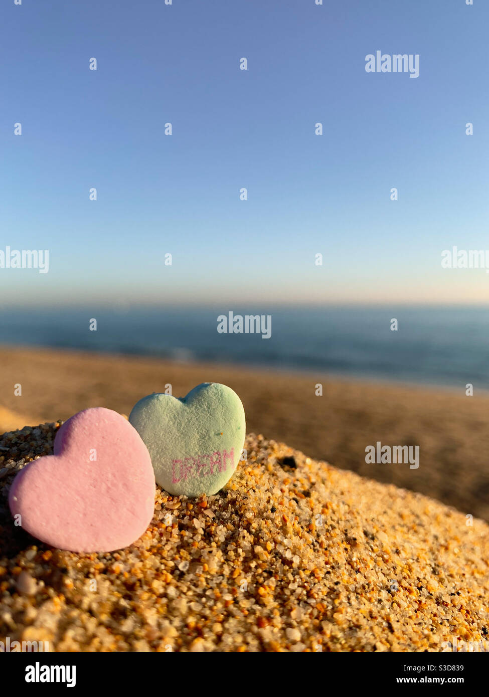 Two heart shaped Valentine candies in the sand on the beach with ocean blurred on the horizon. One of the heart candies has the word dream on it. - Smartphone Captured Stock Image