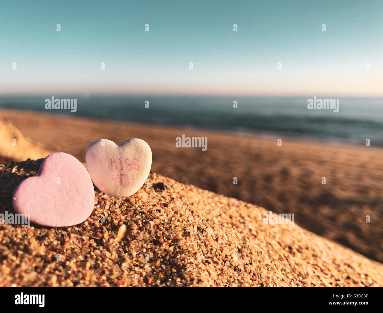 Two Valentine candies in the sand on a sunny beach. One of the candies has the words miss you printed on. - Smartphone Captured Stock Image