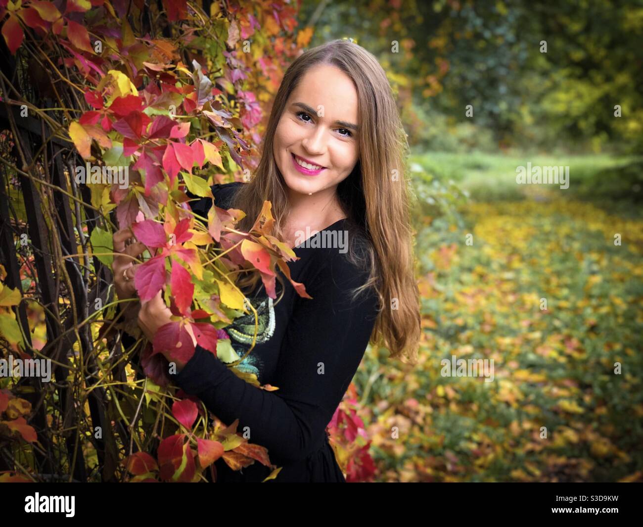 Portrait of happy young woman in black dress standing near a fence covered in colorful ivy leaves - Smartphone Captured Stock Image