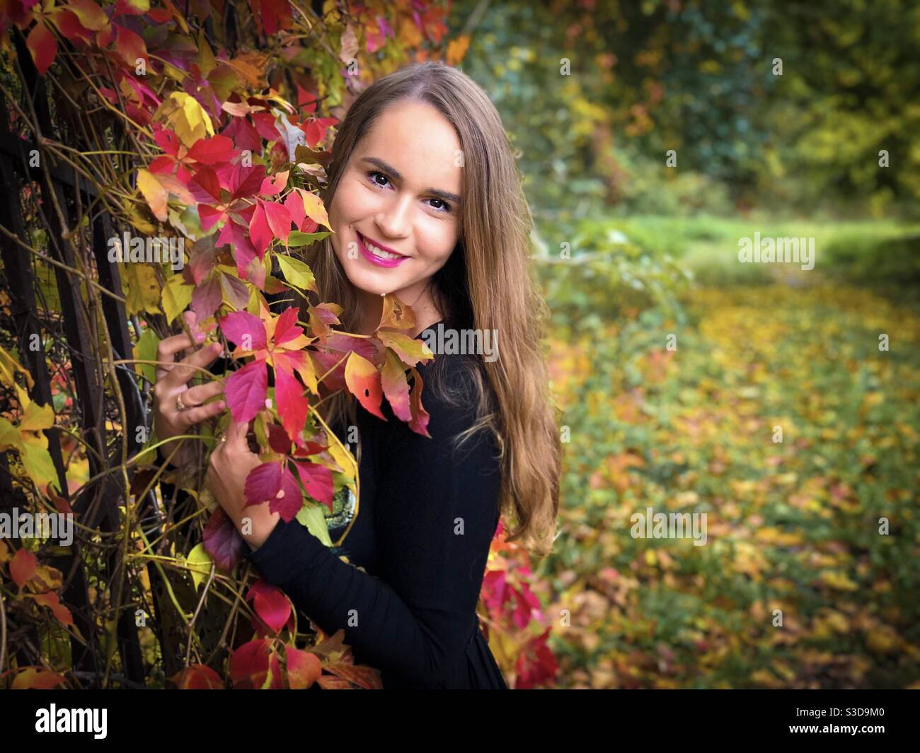 Portrait of happy young woman in black dress standing near a fence covered in colorful ivy leaves - Smartphone Captured Stock Image