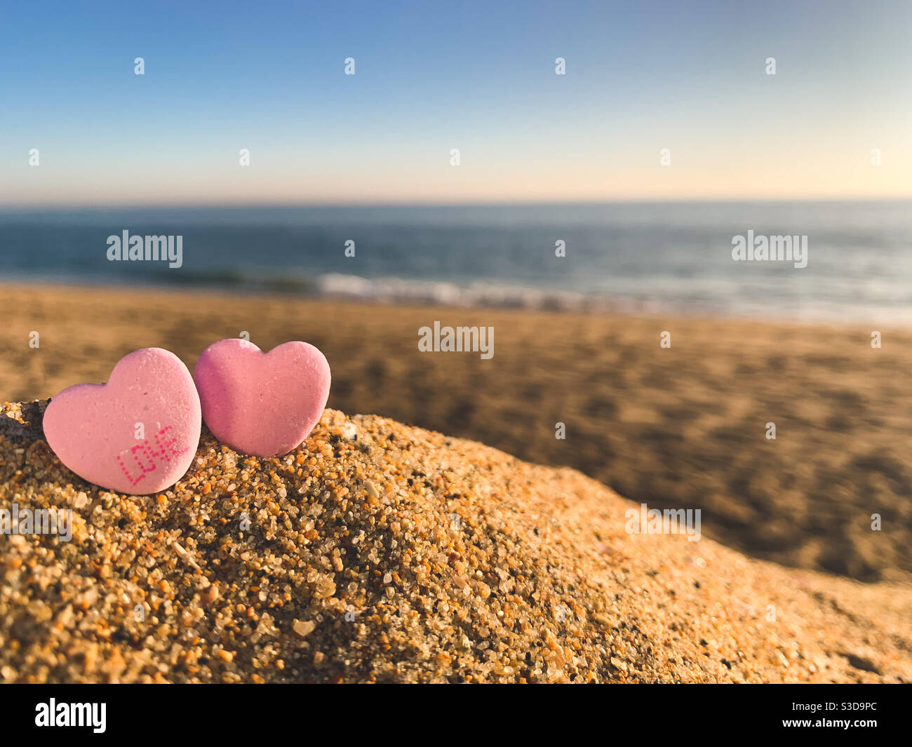 Two pink candy hearts, one with the word “love” on it, sitting in the sand on a sunny beach with ocean blurred in the distance. Stock Photo