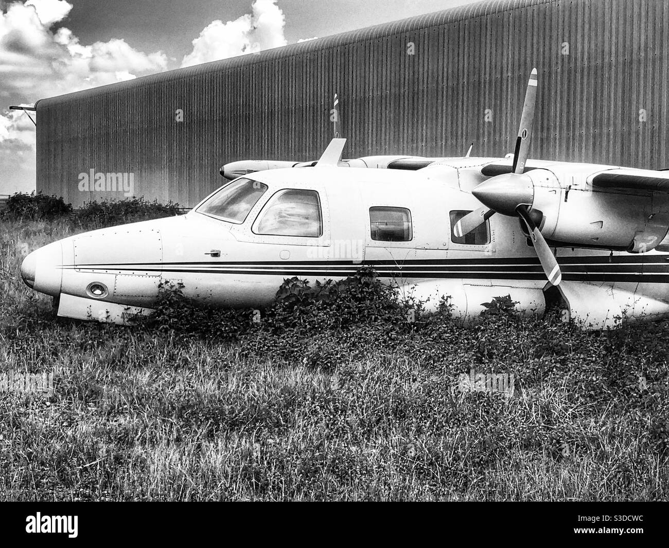 Black and white photo of an abandoned aeroplane lying in long grass outside a hangar at an airfield - Smartphone Captured Stock Image