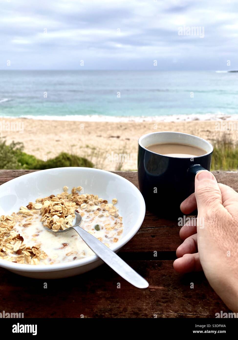 Person enjoys a scenic coastal beach view on a cloudy morning over while having a coffee and breakfast muesli. Concept lifestyle, travel tranquility and recreation. Stock Photo