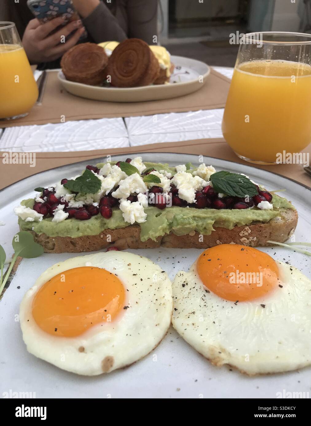Creamy avocado on sourdough bread topped with pomegranate seeds and crumbled feta cheese served with two medium fried eggs and a glass of freshly squeezed orange juice - Smartphone Captured Stock Image