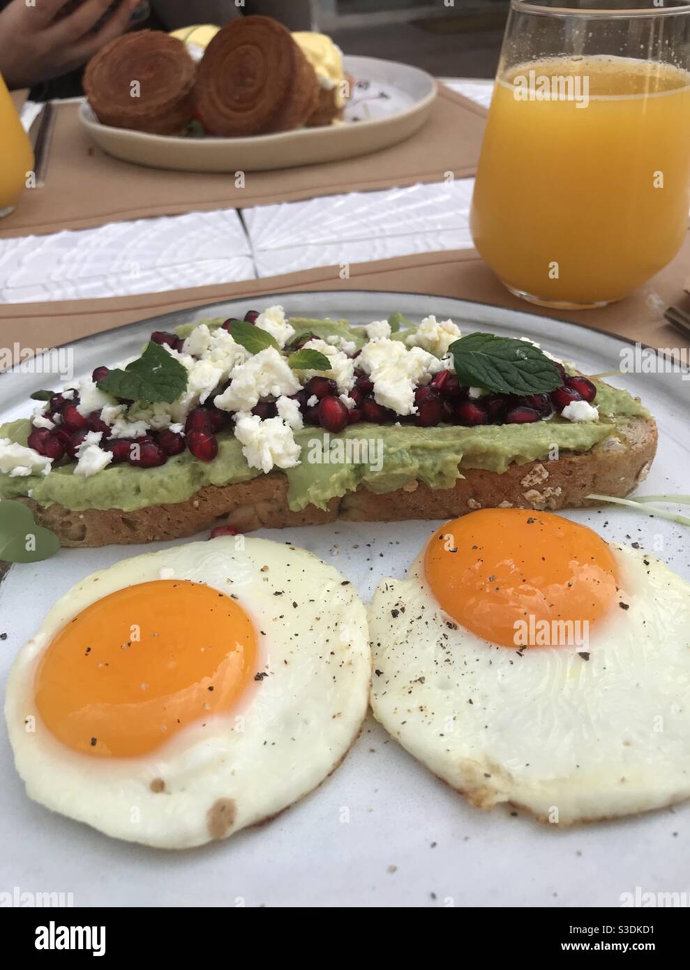 Creamy avocado on sourdough bread topped with pomegranate seeds and crumbled feta cheese and two medium fried eggs served with fresh orange juice - Smartphone Captured Stock Image