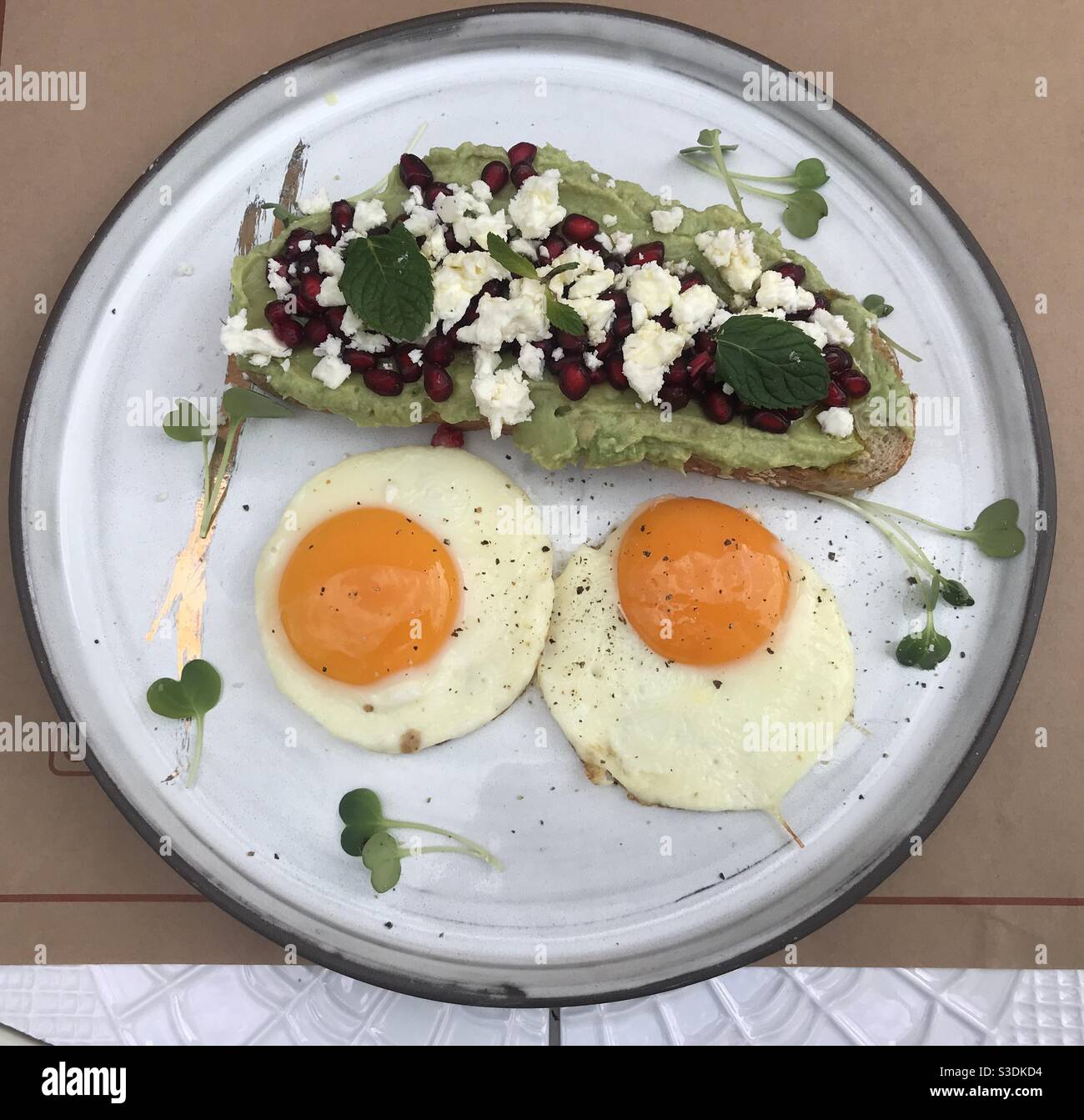 Creamy avocado on sourdough bread topped with pomegranate seeds and crumbled feta cheese and two medium fried eggs - Smartphone Captured Stock Image