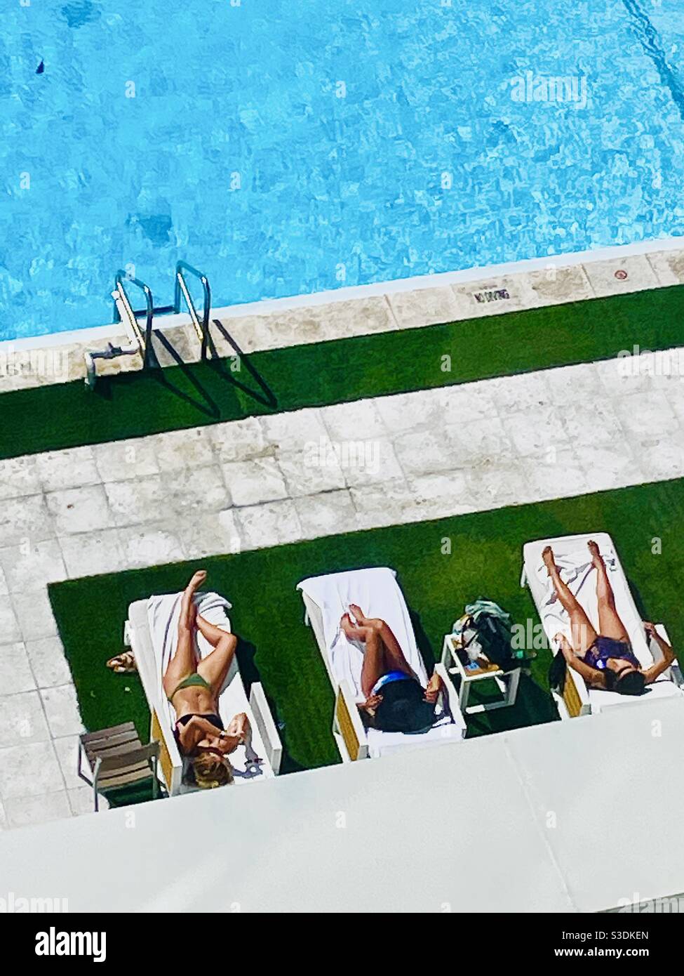 Three young bikini-clad women sunbathing on white lounge chairs beside turquoise water & green astroturf  at the Hilton downtown Miami, 1601 Biscayne Blvd., 33132, USA - Smartphone Captured Stock Image