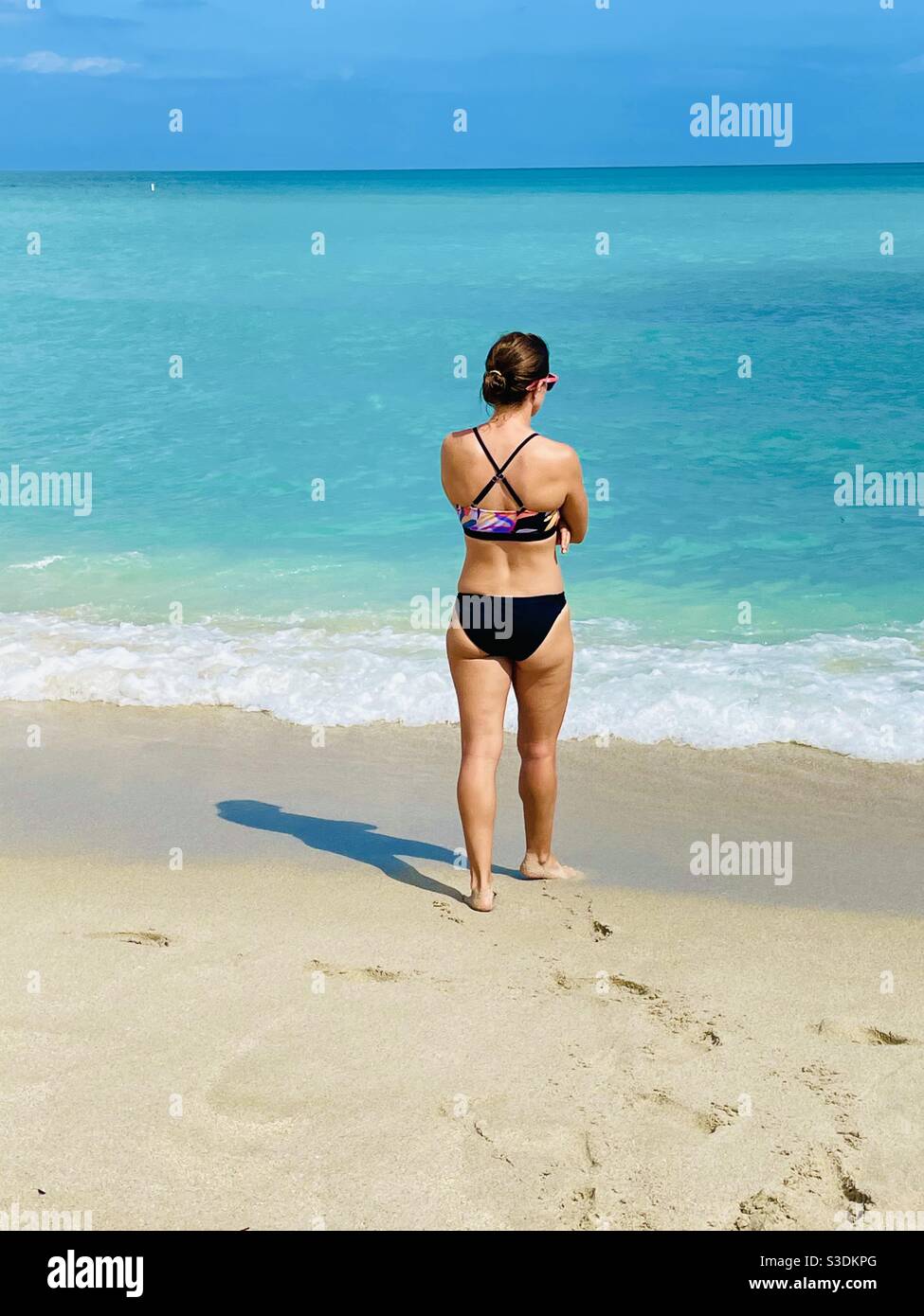 A young brunette woman in a black bikini walks from the sandy beach into the white seafoam at the edge of turquoise ocean water at Miami South Beach, Florid, USA - Smartphone Captured Stock Image