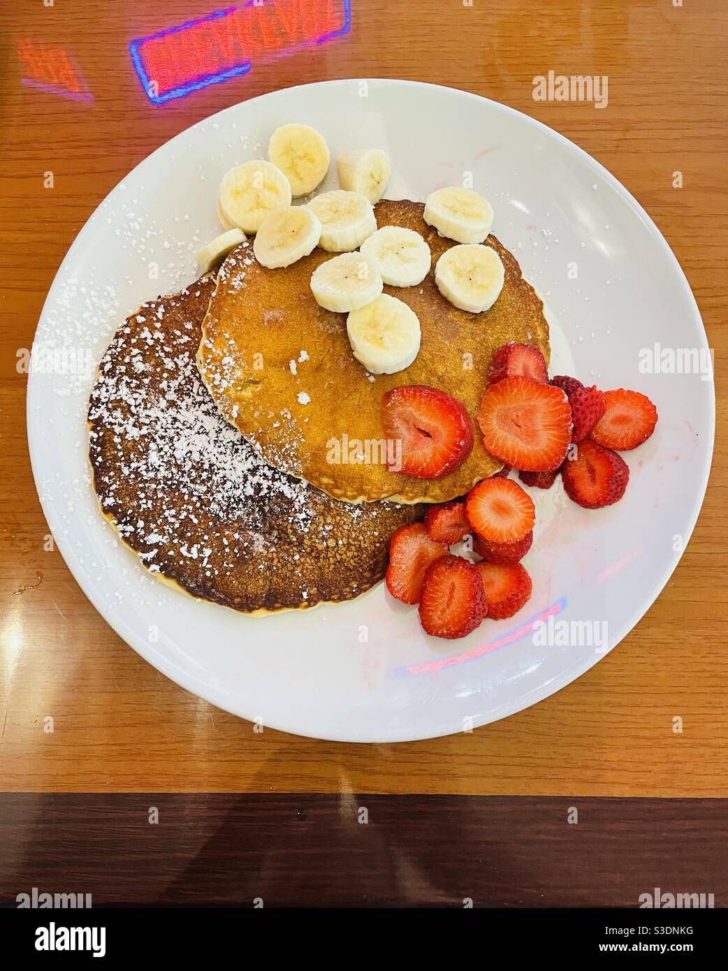 And inviting plate of powdered sugar covered pancakes with sliced bananas and strawberries on a white plate set at a tan-colored table reflecting backwards a red neon sign saying breakfast - Smartphone Captured Stock Image