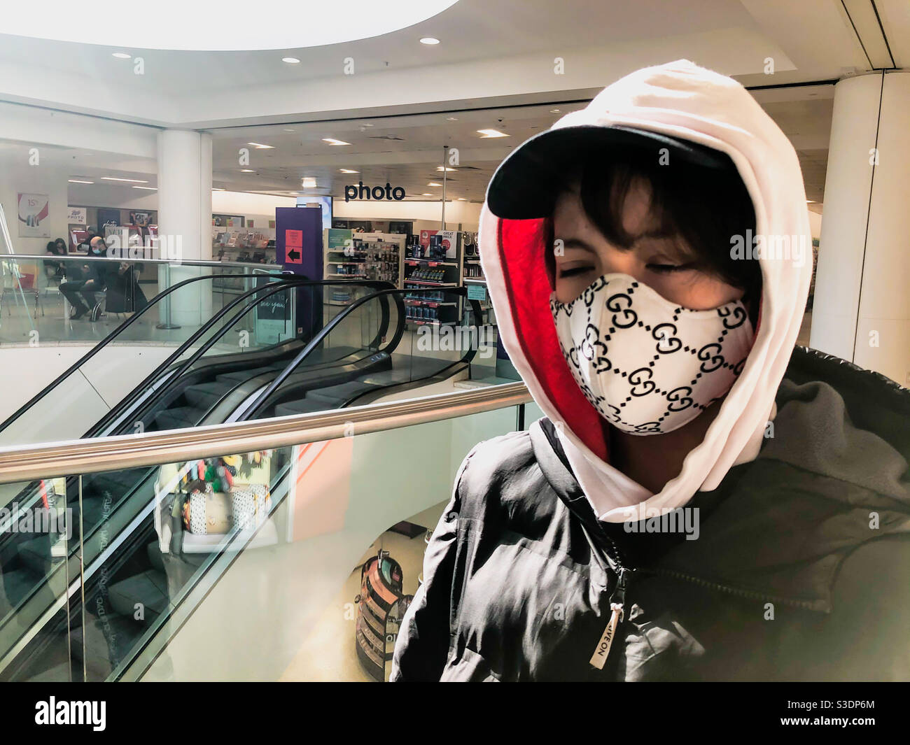 Portrait of a boy wearing a hoodie and a mask next to an escalator in a shop. - Smartphone Captured Stock Image