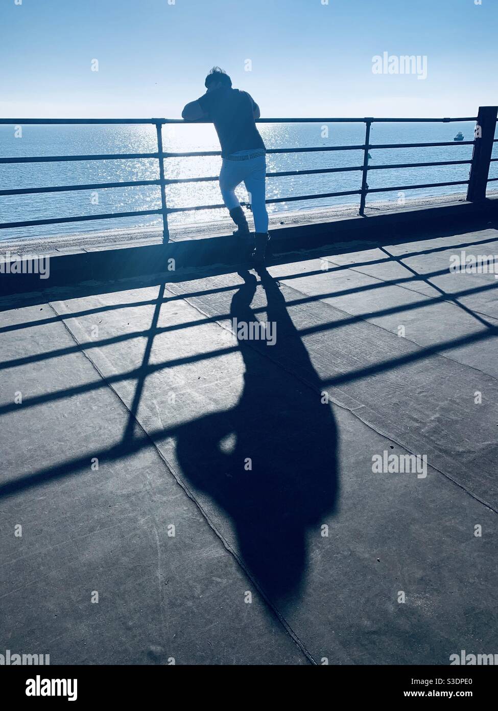 Silhouette of man leaning against railing with sea in background and long shadow Stock Photo