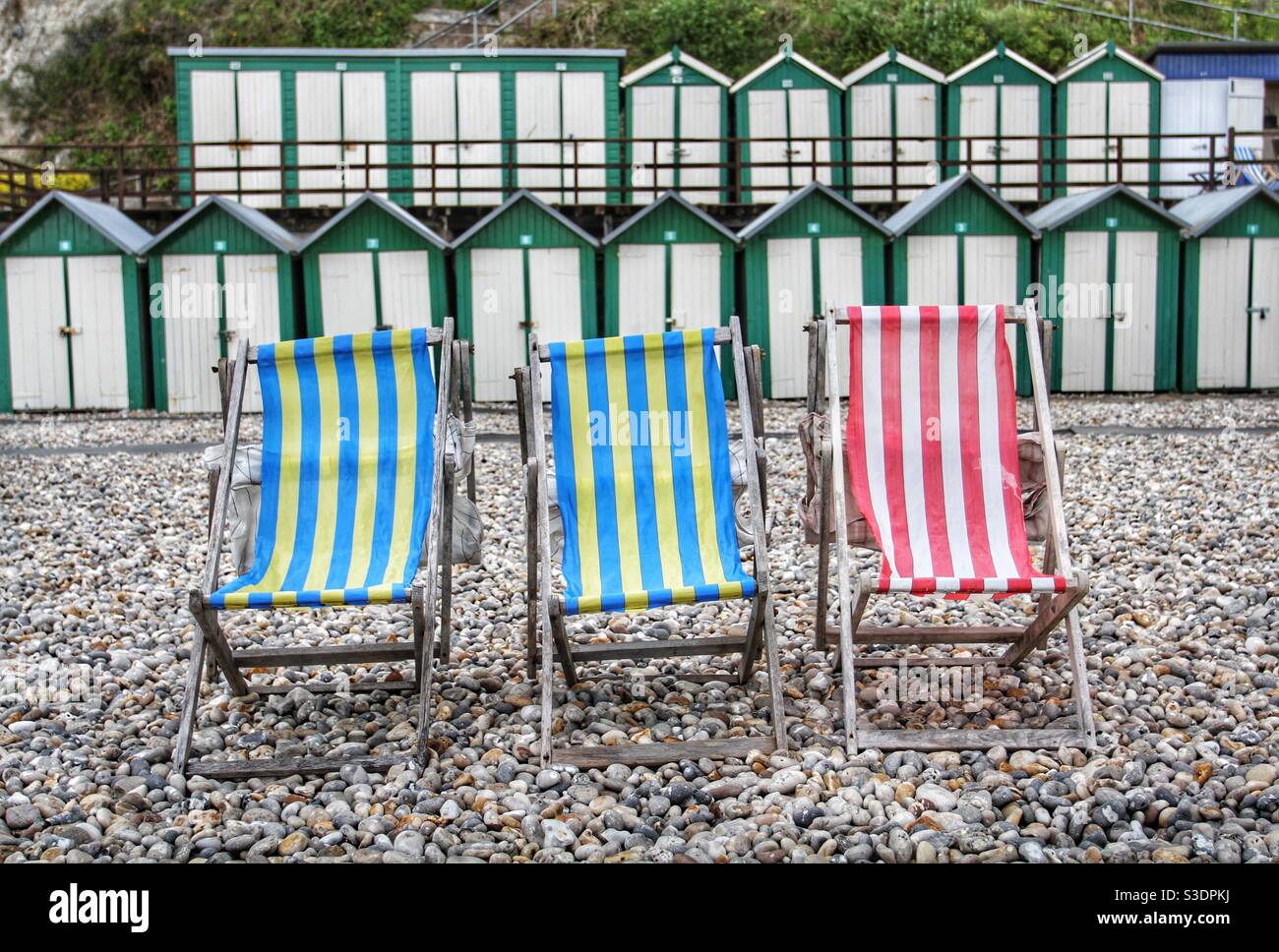 Three traditional stripy deckchairs on a pebble beach at the seaside surrounded by beach huts in the UK - Smartphone Captured Stock Image