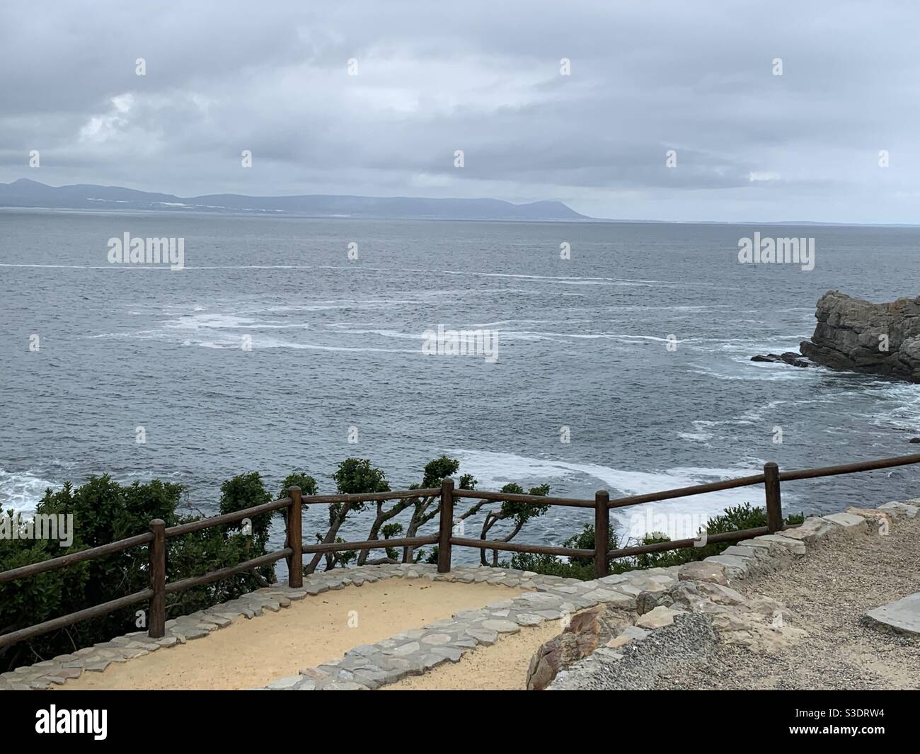 Looking from the shoreline walk in Hermanus across the bay towards Gansbaai. On the near shore we see the walkway, railing and scrub brush above the sea foam. Stock Photo