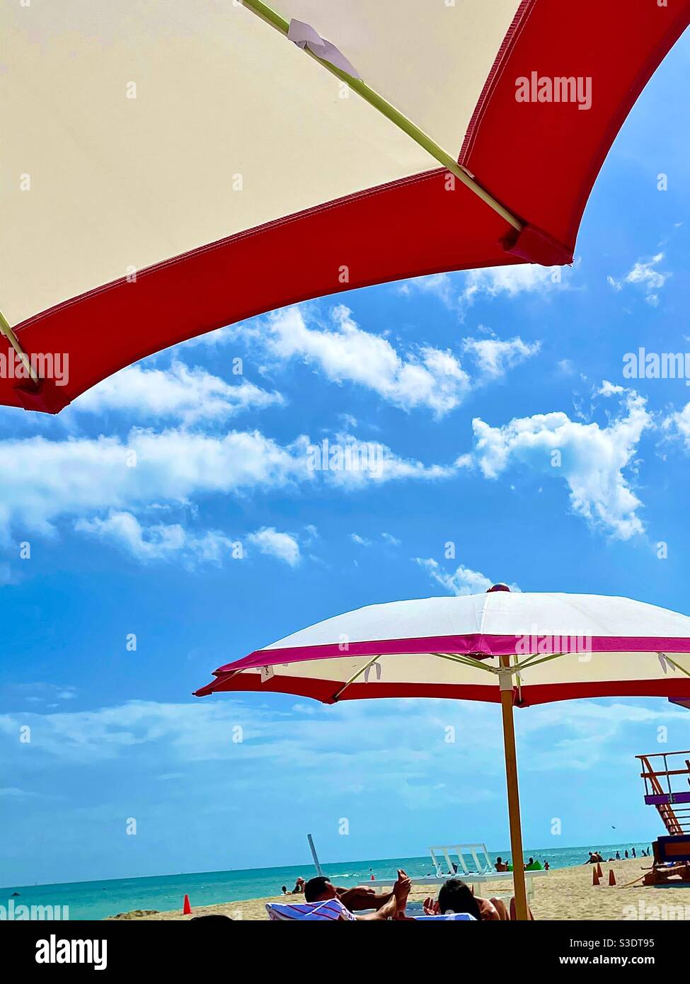 Bright pink trimmed umbrellas help shade beach goers and sunbathers at South Miami Beach, Florida, USA at the oceans edge against  a clear blue sky and white fluffy clouds - Smartphone Captured Stock Image