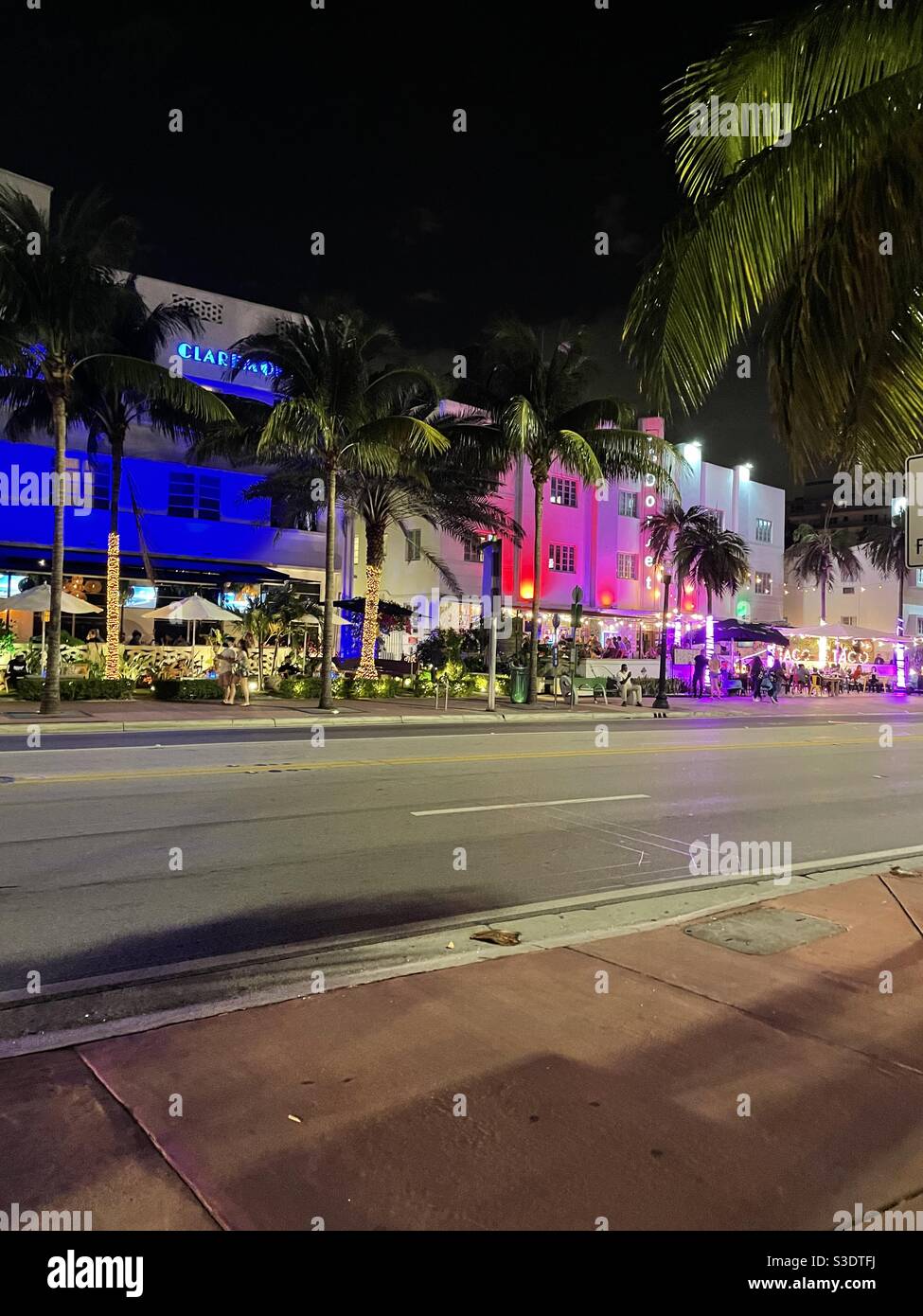 Pink and blue neon Lights glam up the street scene on the popular Collins Avenue Of south Miami Beach, Florida, USA filled with restaurants, shops, outdoor dining, palm trees and people watching - Smartphone Captured Stock Image