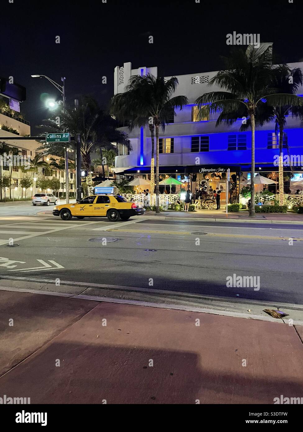 A taxi drives past the Hampton Inn on popular Collins Avenue in South Miami Beach, Florida, USA at night, lit up with blue neon lighting & Has outdoor dining under palm trees and colorful umbrellas. - Smartphone Captured Stock Image