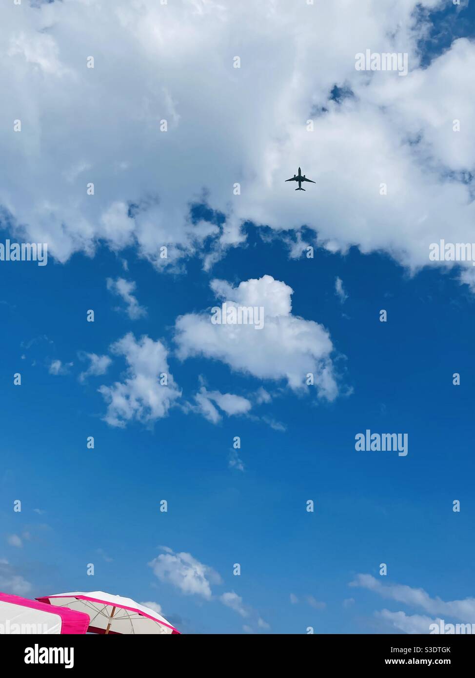 A jet airplane is silhouetted against fluffy white clouds in a blue sky over pink and White umbrellas in Miami South Beach, Florida, USA - Smartphone Captured Stock Image