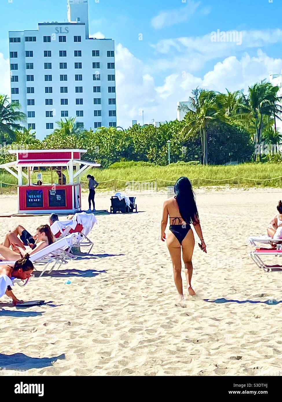 A woman in a black bikini with long dark hair walks past sunbathers on lounge chairs in the sand toward the pink towel shack of the SLS a hotel in Miami South Beach, Florida, USA, 33139 - Smartphone Captured Stock Image