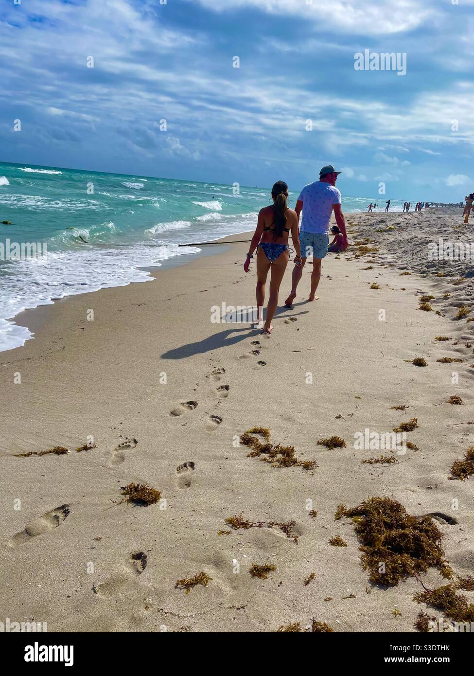 A man and a woman leave foot prints in the sand as they walk on the beach beside whitecapped waves on the turquoise ocean at Miami South Beach, Florida, USA - Smartphone Captured Stock Image