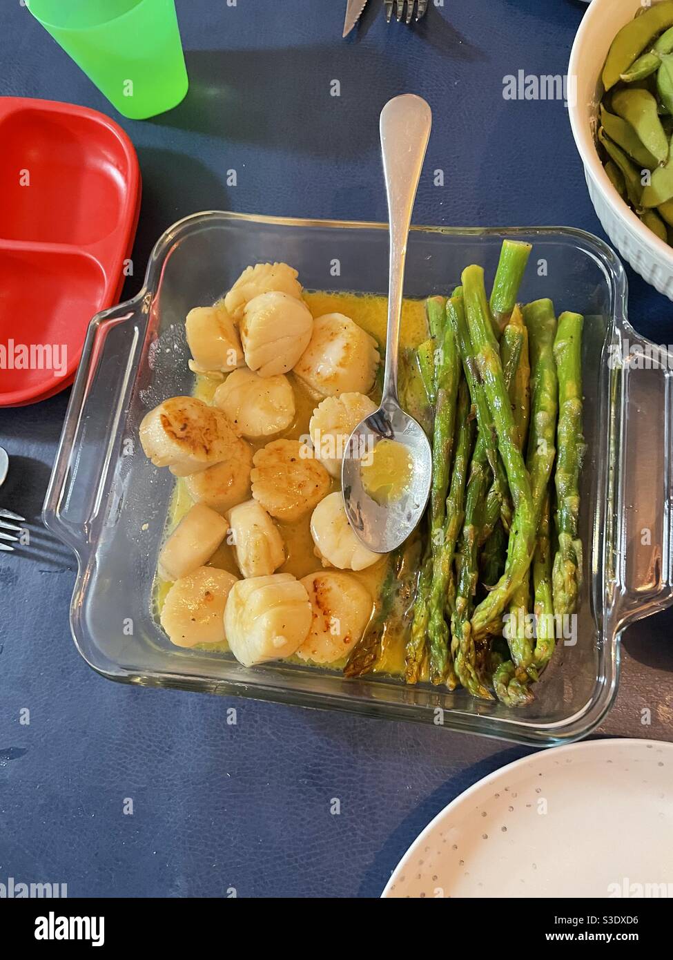 Home cooked scallops and asparagus in a garlicky butter sauce in a glass serving dish With a spoon set on a dark blue tablecloth - Smartphone Captured Stock Image