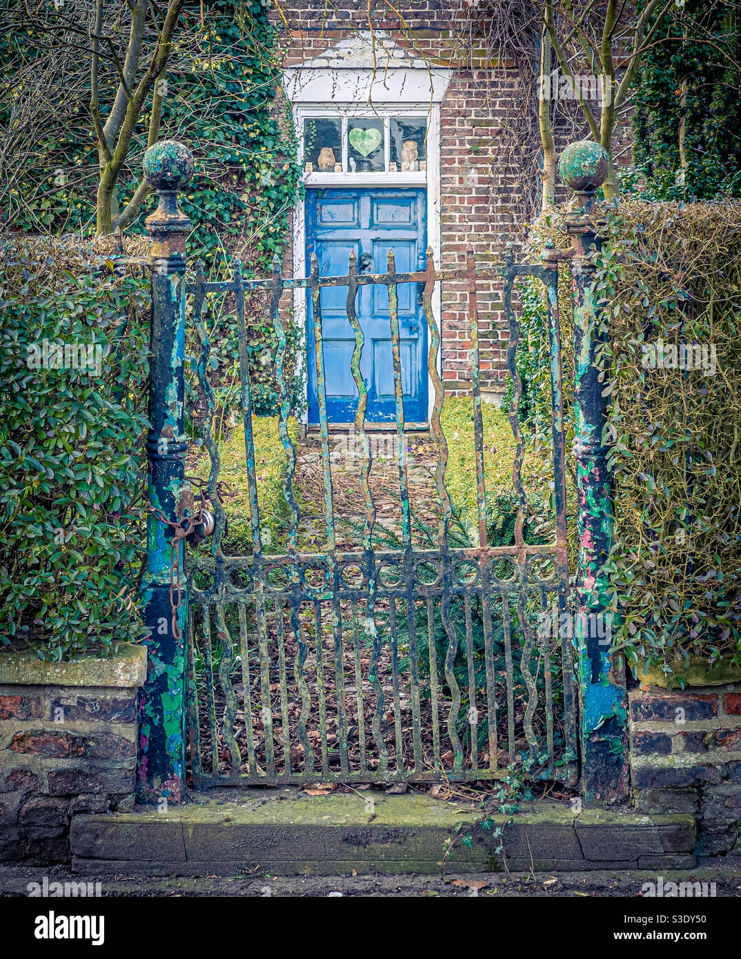 Old wrought iron gate with flaky layers of coloured paint and house in background with blue door - Smartphone Captured Stock Image