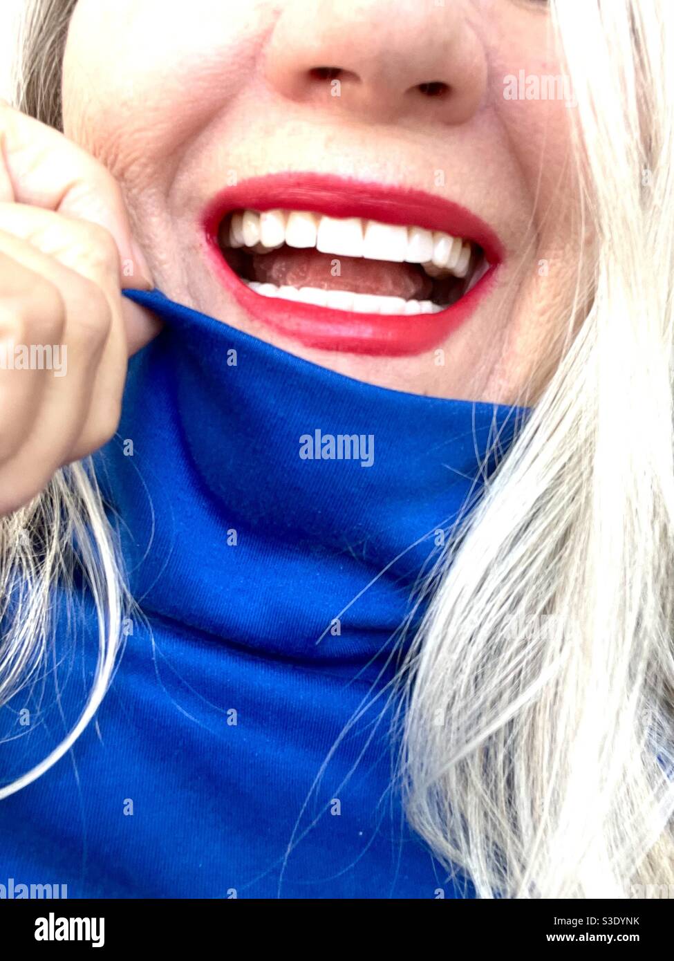 A bright toothy smile with red lipstick against a cobalt blue turtleneck and pale blonde hair - Smartphone Captured Stock Image
