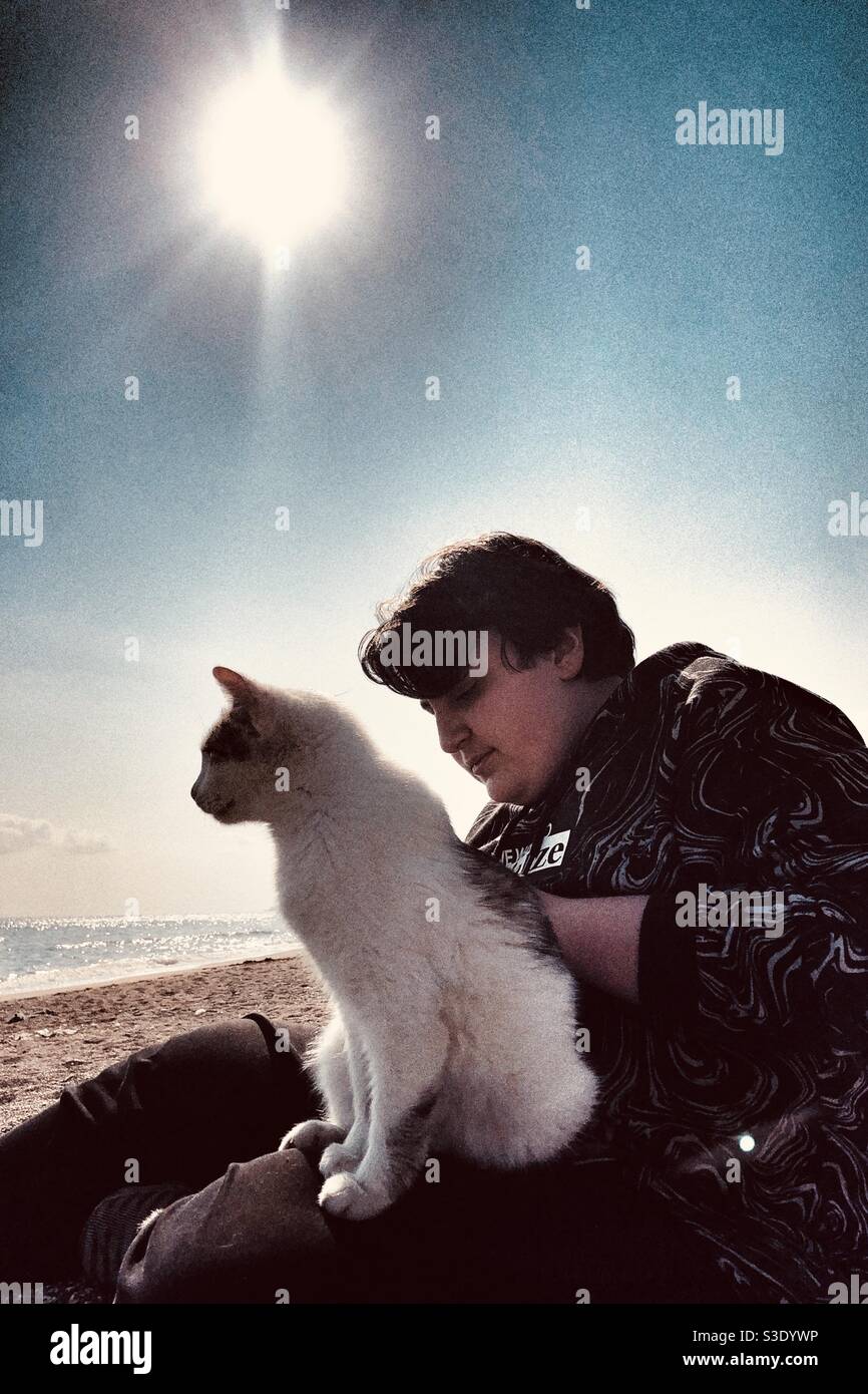Teenage boy sitting on a beach with a cat strutting on the lap Stock Photo