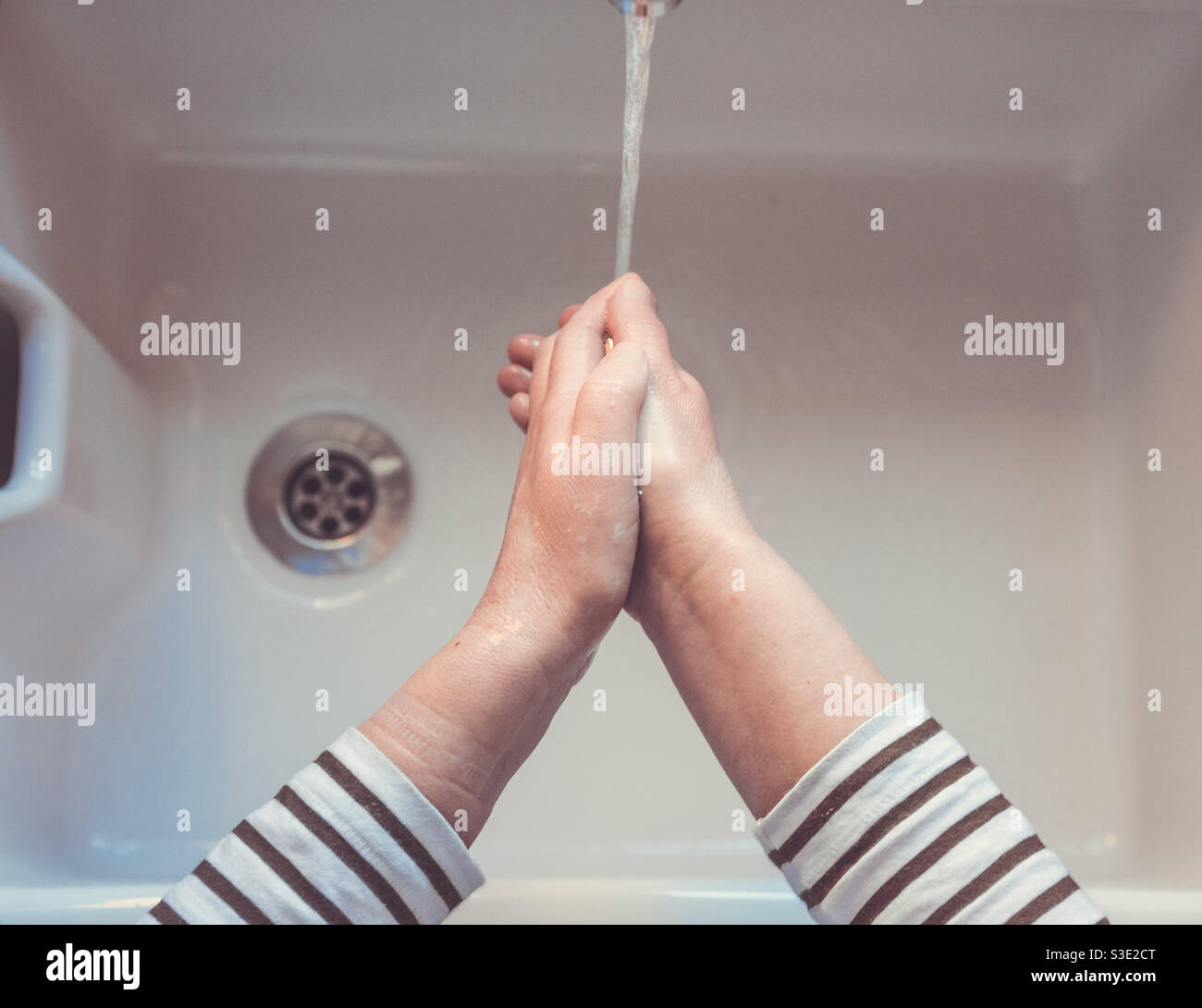 Point of view of a pair of hands washing under a flowing tap and running water above a sink with plug hole Stock Photo