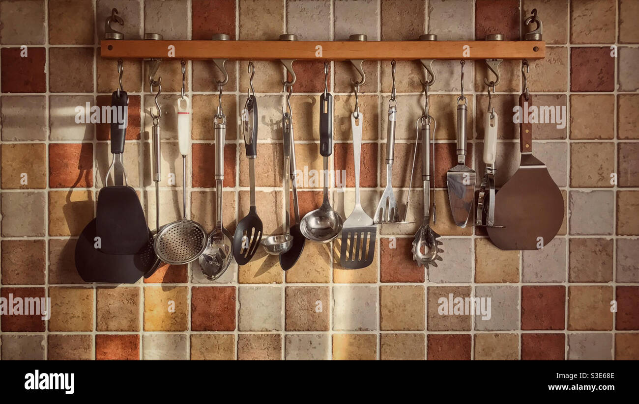 Rack of hanging kitchen utensils against a terracotta tiled wall. Stock Photo