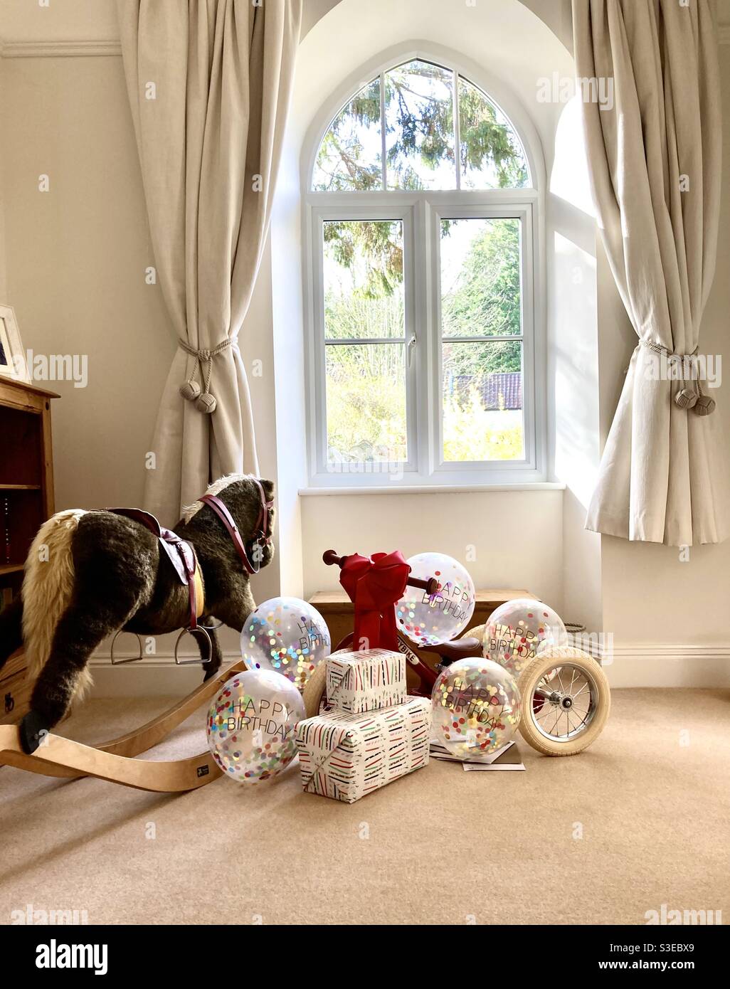 First birthday presents (bike and rocking horse) and balloons by the window Stock Photo