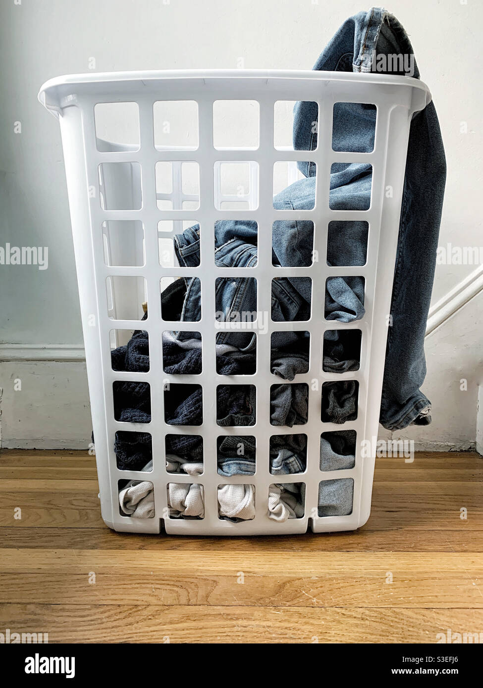 Laundry in white plastic basket near laundry room, a pair of jeans hanging over side - Smartphone Captured Stock Image