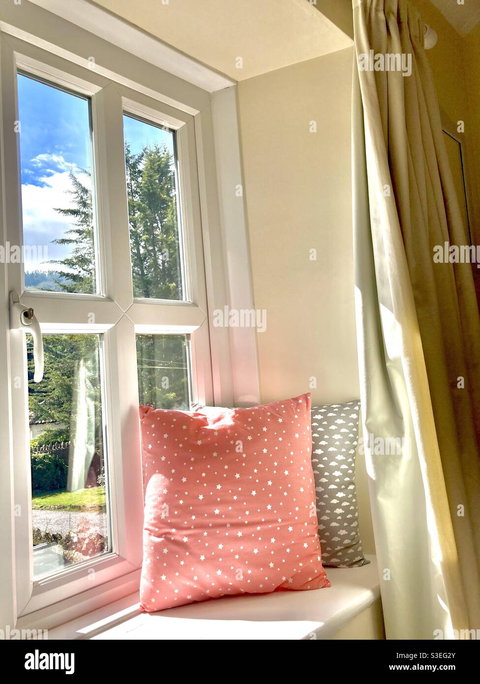 Window seat in the sun with coral and grey cushions and cream curtains. Stock Photo